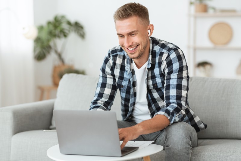 Homme souriant travaillant sur un ordinateur portable depuis son canapé, en tenue décontractée.